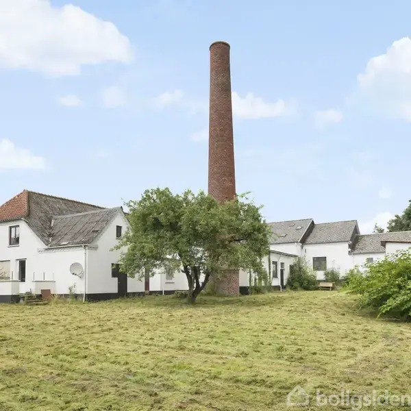 En hvid, landlig bygning med rødt tegltag står ved siden af en høj, rød murstensskorsten. Græsplæne og træer omgiver bygningen under en skyet himmel.