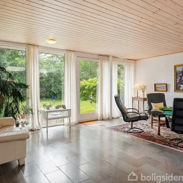 En stue indeholder en hvid sofa og sorte stole i samtaleområde. Store vinduer viser en have udenfor. Et maleri hænger på væggen. Gulvtæppe og planter skaber en hyggelig atmosfære.