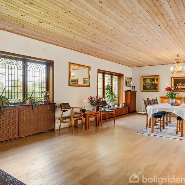 Et rummeligt spiseområde indeholder et rundt bord dækket med hvid dug og omgivet af stole. Trægulve og loft tilføjer varme, mens vinduer med blyindfattede ruder lader lys strømme ind.