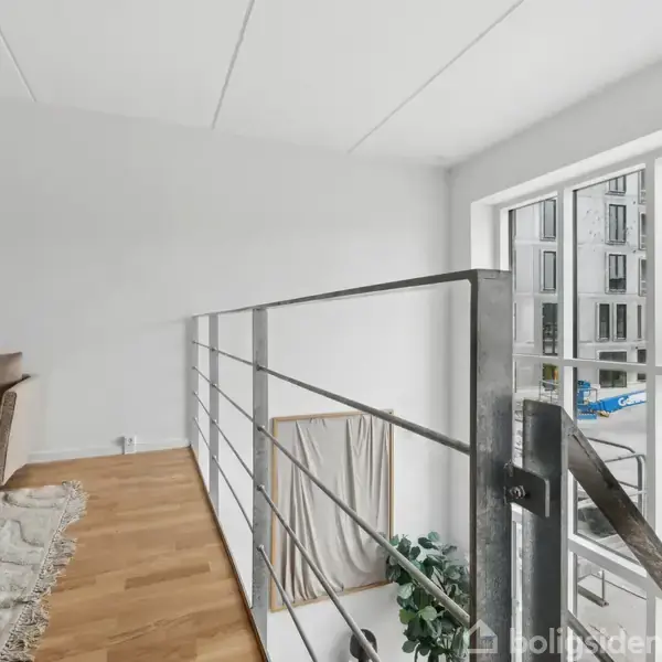 Sofa står ved siden af et balustrade på en hems. Store vinduer giver udsigt til moderne lejlighedskompleks udenfor. Trægulv og tæppe skaber en hyggelig atmosfære.