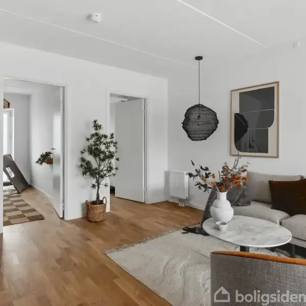Stue med grå sofa og rundt marmorbord, dekoreret med vaser. Rummeligt trægulv, hvide vægge, moderne lamper og døråbninger til andre værelser, giver et lyst, indbydende miljø.