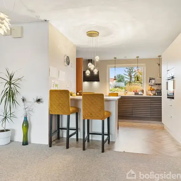 To gule barstole står ved en køkkenø i et lyst og moderne køkken med hængende lamper og planter, mens en åben dør viser en garderobe.