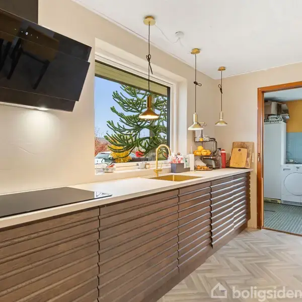 Køkken med brunstribet skab, sort emhætte, moderne kogeplade og vindue. Hængelamper over vask. Til højre, dør til vaskerum med vaskemaskine og tørretumbler.