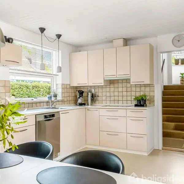 Køkken med lyse skabe og opvaskemaskine, i brug med kaffemaskine og planter. Spisebord med blomster i forgrunden, trappe op til en anden etage. Ingen tekst fundet.