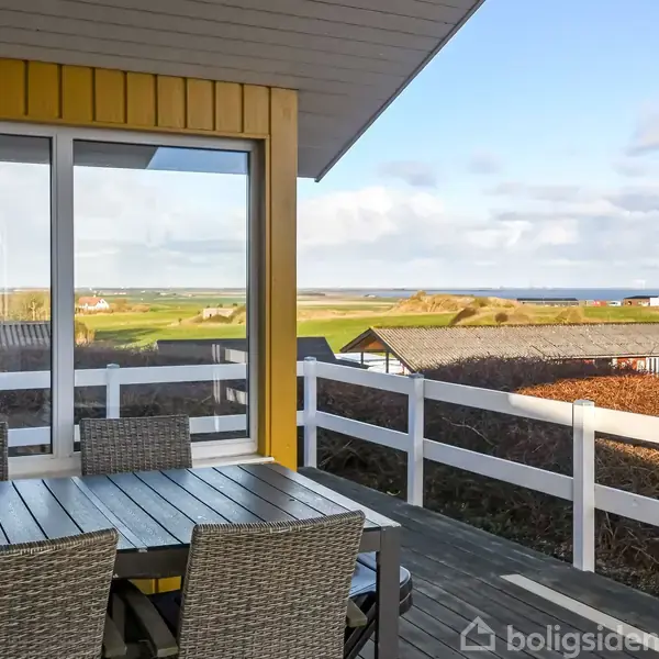 Terrasse med træbord og flettede stole i forgrunden. Udsigt over landskab med græsarealer, spredte huse og hav i baggrunden. Skyet himmel giver en overdøvet belysning.