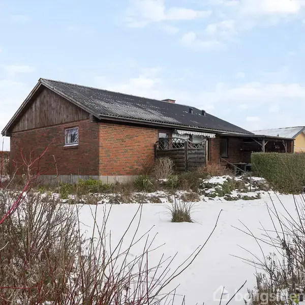 Et rødt murstenshus står stille i en snedækket have med buskads omkring. Taget er mørkt, og der er en træveranda. Himlen er let overskyet.