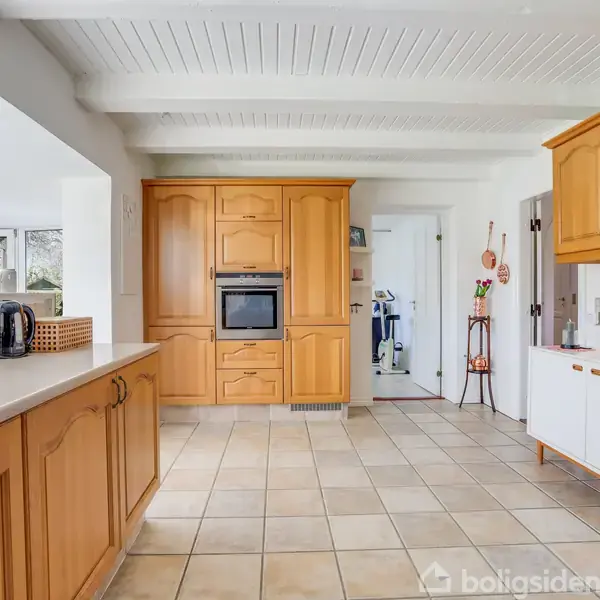 Køkken med træskabe og indbygget ovn, flisegulv og hvide vægge. En elkedel og brødrister står på bordpladen, og der er et lille hjørne med udgang til en anden del af huset.
