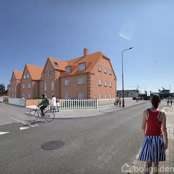 Et stort rødt murstenshus med flere kviste og orange tegltag står ved et vejkryds. En cyklist passerer, mens folk går forbi. Solrig dag og blå himmel.