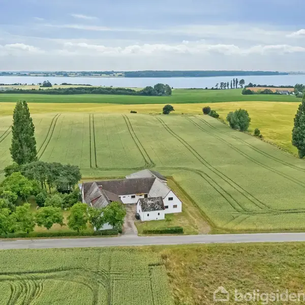Et hvidt bondehus med stråtag ligger stille mellem grønne marker. Omkring huset er der træer. I baggrunden ses en vej og en fjord eller sø i horisonten.