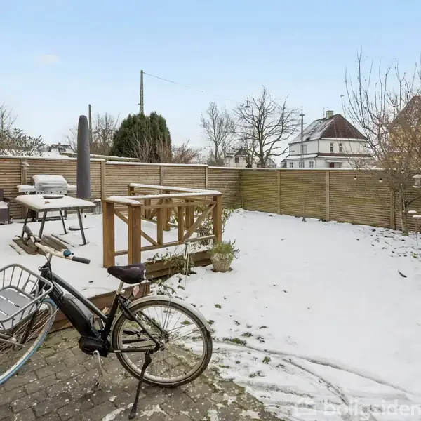 Cykel står stille på snedækket terrasse med stole og grill i baggrunden, omgivet af træhegn og huse i et beboelsesområde.