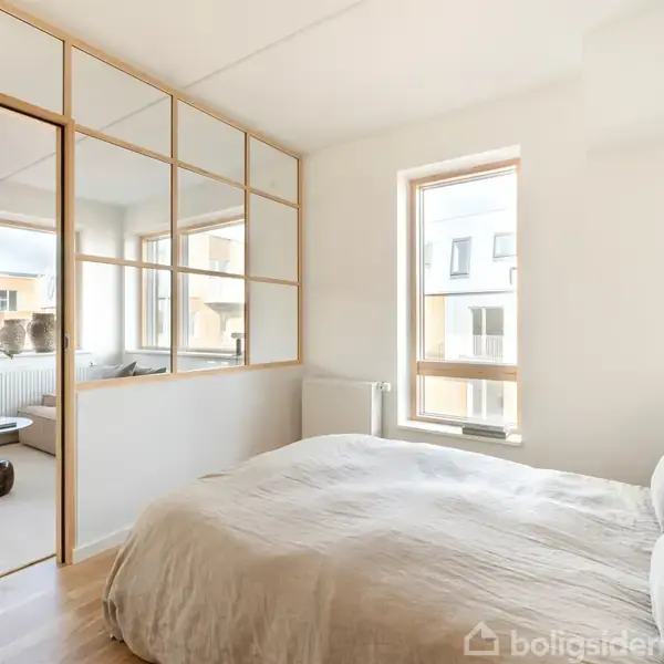 Soveværelse med stor seng dækket af lysegråt sengetøj. Ved siden af sengen står en lampe, og rummet har store vinduer med en udsigt til nabobygninger. Skydedøre adskiller rummet fra en stue med moderne indretning.