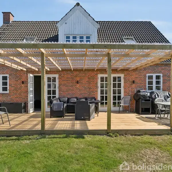 Et rødstenshus med trekanter på taget. Foran huset er en stor træterrasse med hvide møbler og en grill. Terrassen er indrammet af en pergola. Græsplæne omgiver terrassen.