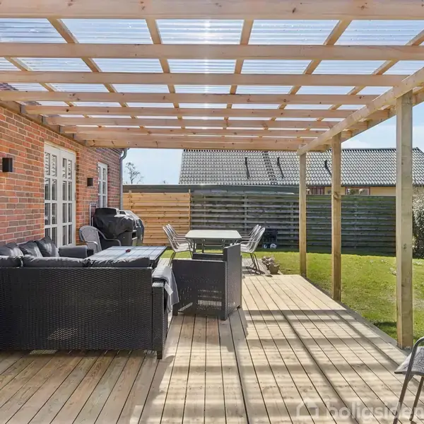 En overdækket træterrasse med sorte havemøbler og grill står ved et rødt murstenshus. Græsplæne og hegn omkranser området i baggrunden.