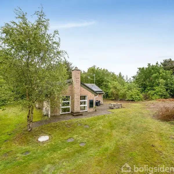 Et lille hus med en skorsten står i en grøn have omgivet af træer og buske. Huset har flere vinduer og en terrasse med et bord og bænke.