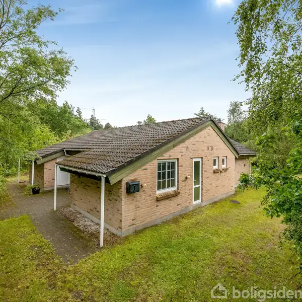 Et murstenshus med et mørkt tegltag står omgivet af frodig vegetation. Huset har en hvid dør og vinduer, og en grusvej leder til indgangen. Atmosfæren er fredelig og grøntfyldt.