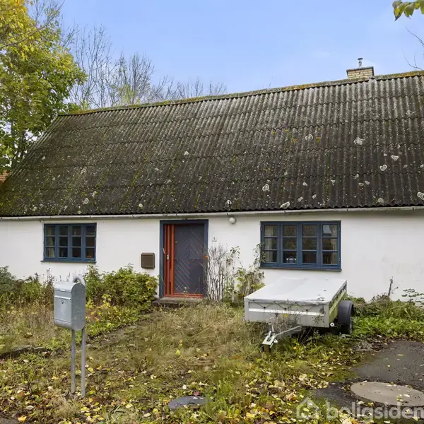 Hvidt hus med skråt tag og to blå vinduer omkring en centralt placeret dør. En trailer står parkeret ved siden af. Omgivet af højt græs og træer i en villavej.
