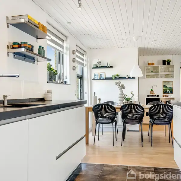 Køkken med hvide skabe og sort bordplade. Fire sorte stole står ved spisebordet. Hylder med planter og kogebøger hænger på væggen. Lysindfald gennem store vinduer.