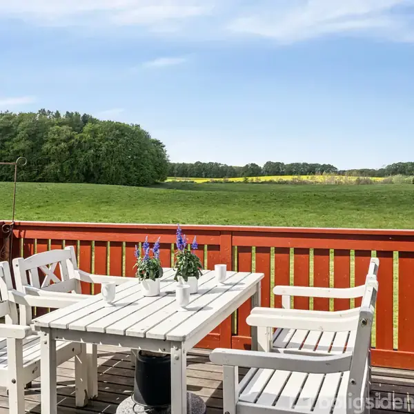 Et hvidt havebord med fire stole står på en terrasse med rødmalet rækværk. Bordet har potteplanter, og terrassen har udsigt over grønne marker og skov i baggrunden.