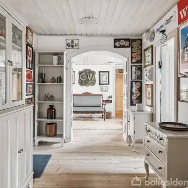 En lys gang med hvide skabe og hylder viser forskellige dekorationer og skilte. Midt i billedet ses en buegang ind til en stue med et træmøbleret bord. Tekst: "J.C.W. Garage".
