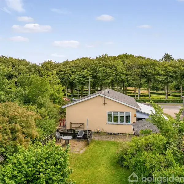 Et gult hus står fredeligt omgivet af frodige træer og buske. Foran huset er der en velplejet græsplæne og terrasse, og der er skov i baggrunden.