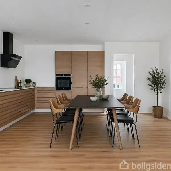 Spisebord med ti stole står i et moderne, lyst køkken-alrum med trægulv. Køkkenet har trækabinetter, indbygget ovn, planter, og minimalistisk dekoration, inklusiv et abstrakt maleri og en lanterne.