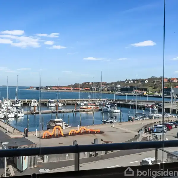 Havn med flere fortøjede både; biler parkeret ved kaj. I baggrunden ses huse på en bakke. Himlen er klar og blå.
