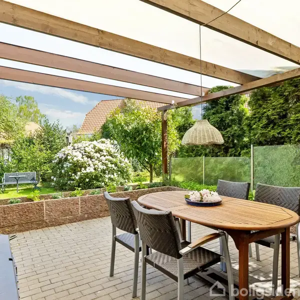 En overdækket terrasse har et rundt bord med fire stole, udsigt til en velplejet have med blomstrende buske, en bænk og frodig grøn vegetation.