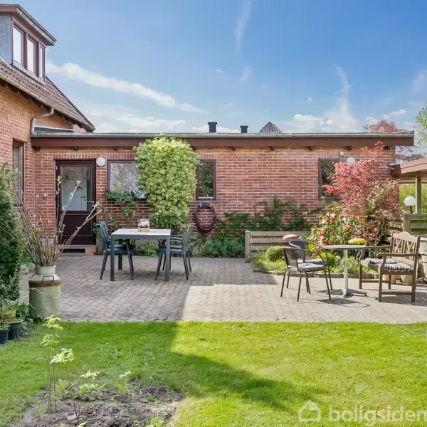 Murstenshus i en baghave med terrasse. Havemøbler står på flisebelagt område omgivet af blomsterbede. Græsplæne strækker sig fremad. Et lille bord og stole ses i højre side.