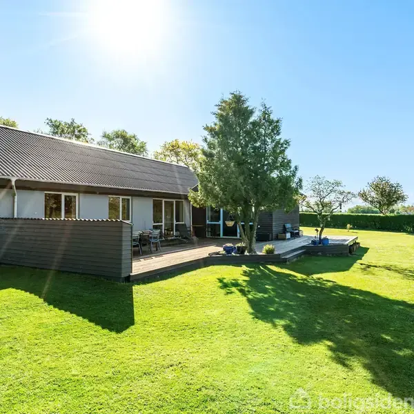 Hus med terrasse står stille i en solrig, velplejet have med grøn plæne og træer. Solen skinner klart på himlen og kaster skarpe skygger.