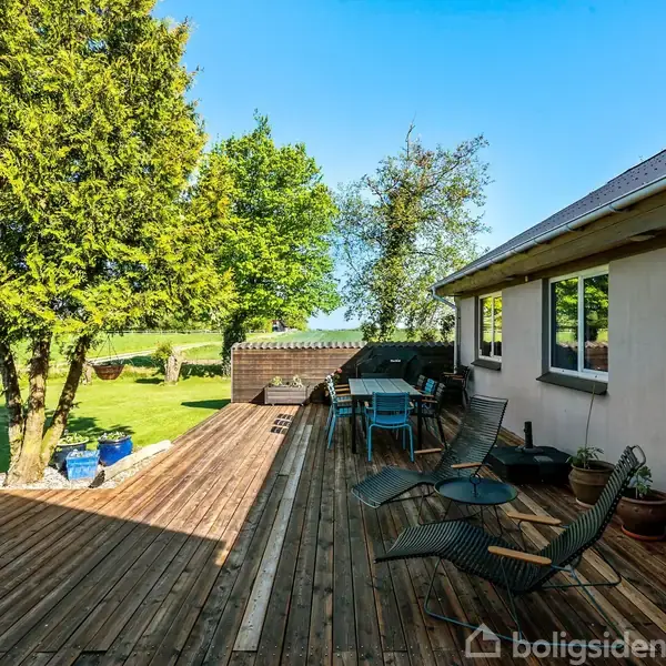 Terrasse med træmøbler og liggestole, solbadning i en frodig have. Huset til højre har store vinduer, og omgivelserne inkluderer græsplæne, træer og en lys blå himmel.