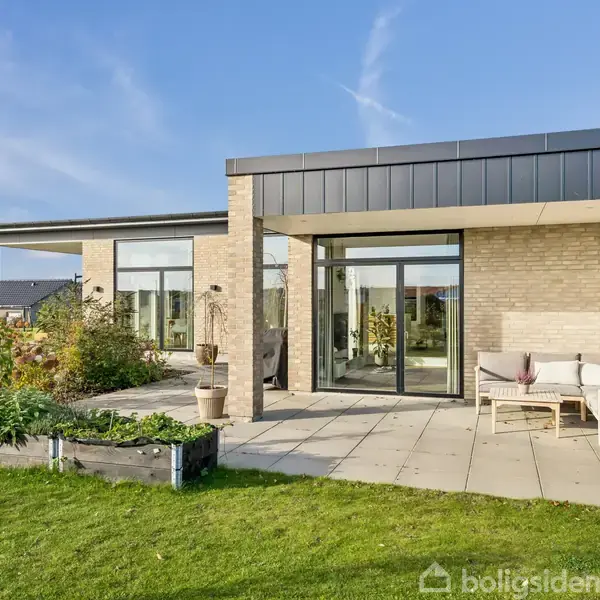 Moderne enfamiliehus med fladt tag og murstensfacade omgivet af en have. Solbeskinnet terrasse foran huset med havemøbler og potteplanter. Klar blå himmel i baggrunden.