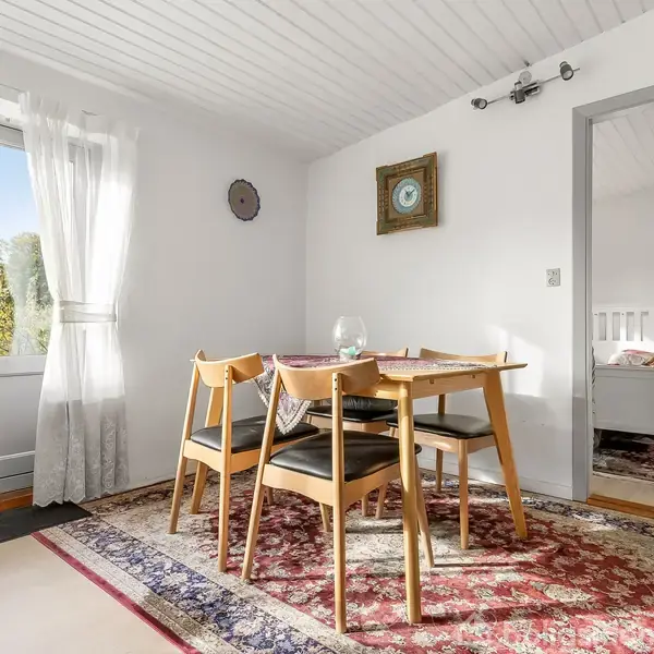 Bord omgivet af fire stole står på et tæppe i en lys spisestue med hvide vægge. En dør fører til en anden del af huset, og vinduer giver naturligt lys.