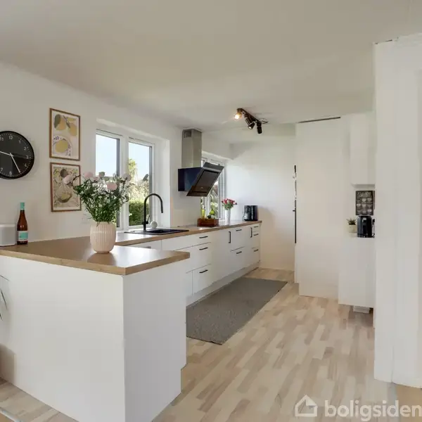 Hvidt køkken med træbordplade står i et rummeligt, lyst rum. En vase med blomster og en vinflaske dekorerer bordet. Køkkenudstyr og vægkunst kompletterer indretningen.