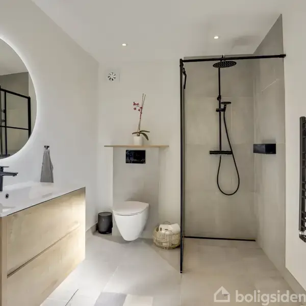 Rundt spejl lyser over et træskab med vask. Brusekabine har glasvæg og sort armatur. En potteplante står ved toilettet. Moderne badeværelse med hvide vægge og håndklædetørrer.