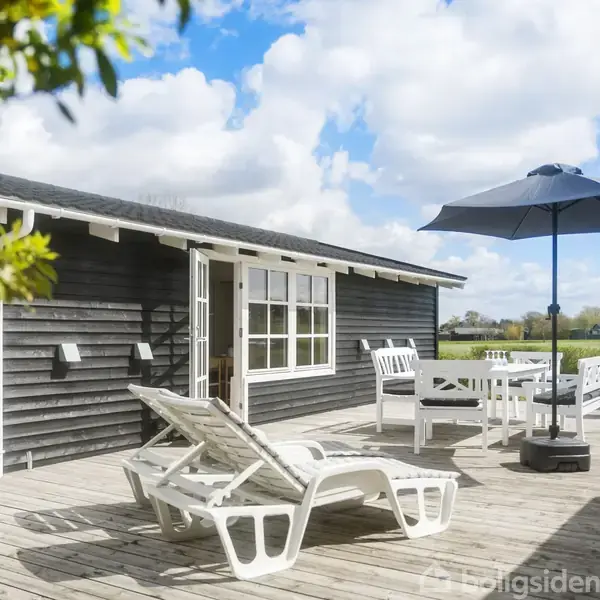 Solstole og bord står på en træterrasse foran et sortmalet sommerhus med store vinduer. En parasol giver skygge, mens blå himmel og skyer danner baggrunden.