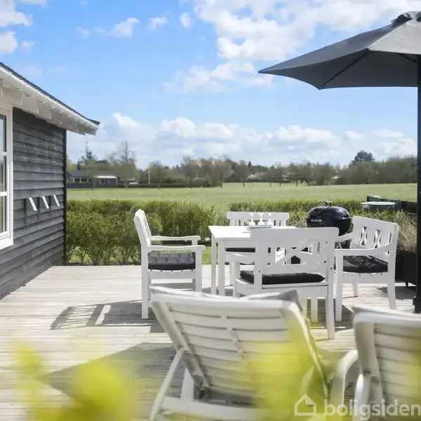 Udendørs terrasse med hvidt bord og stole under sort parasol, ved et sort træhus. Åben dør leder udenfor. I baggrunden ses en grøn mark og træer under blå himmel.