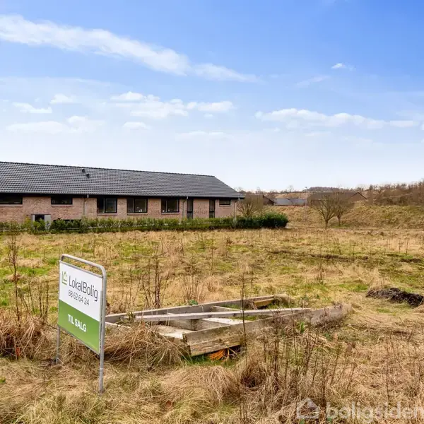 Et skilt fra "Edc LokealBolig" viser "Til Salg" foran en græsmark, med en rækkehusbygning i baggrunden og en blå himmel ovenover.