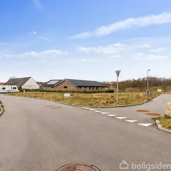 Vej mødes skæringspunktet; roligt boligområde med huse og parkerede autocampere i baggrunden; omgivet af græsarealer og vejskilte; overskyet himmel.