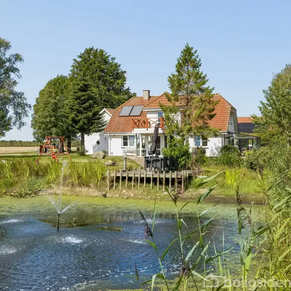 Et hvidt hus med rødt tag står i en frodig have. En lille dam med en fontæne er foran huset, omgivet af træer og græs.