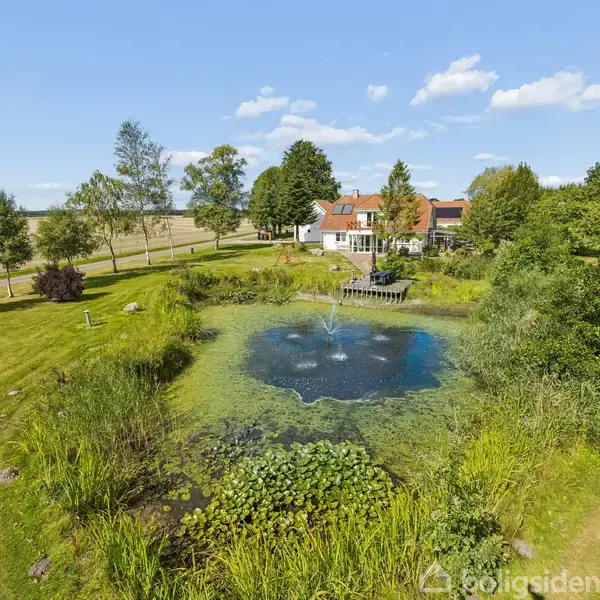 En dam med springvand i aktion omgivet af frodige græsarealer og træer med et hus i baggrunden. Klar blå himmel med få skyer.