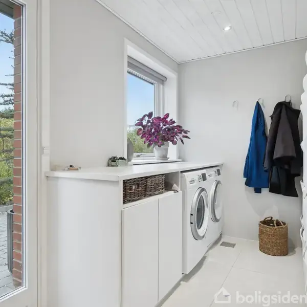 Vaskemaskine og tørretumbler står ved siden af hinanden på gulvet i et lyst rum med hvidt loft og vægge. En dør, garderobestativer, kurv og et vindue omgiver dem. En plante står på bordet.