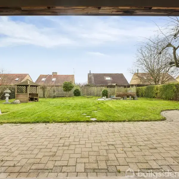 En græsplæne, omkranset af hække, ligger foran en række huse med rødt tegltag. En flisebelagt terrasse ses i forgrunden, og der er et træ til højre.
