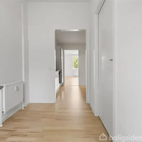 En lang gang med lyse trægulve strækker sig ind til et rum med et vindue for enden. Til venstre er en radiator og til højre ses en lukket dør.