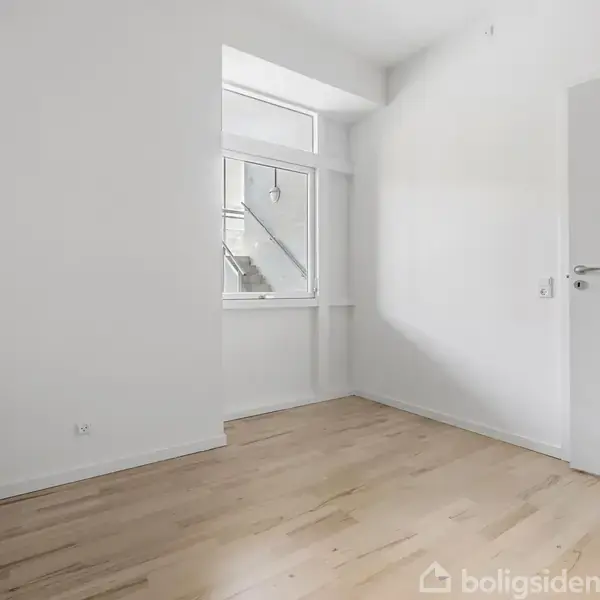 Et tomt, hvidt rum med trægulv; en radiator hænger til venstre. Et stort vindue viser en trappe udenfor. En dør står halvåben mod højre væg.