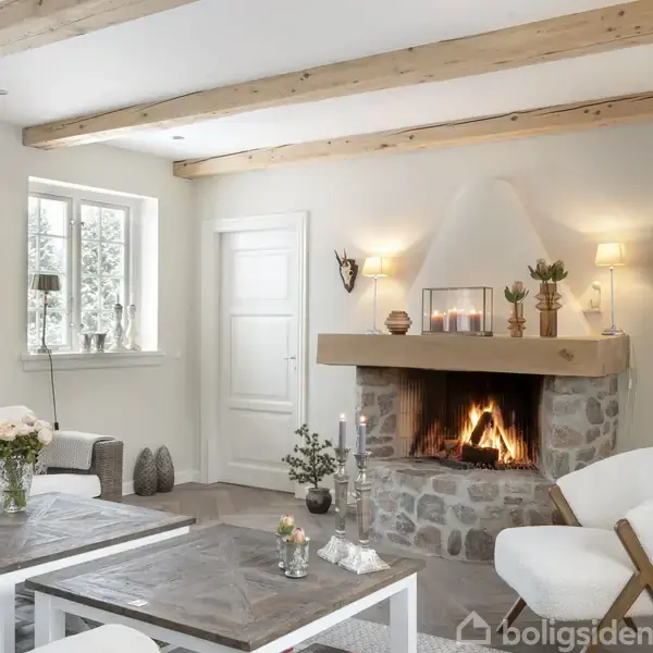 En hyggelig stue med pejs, hvor ilden brænder. Omgivet af hvide vægge, en sofa, et bord med blomster og pynteting på kaminhylden under blødt lys fra væglamper.