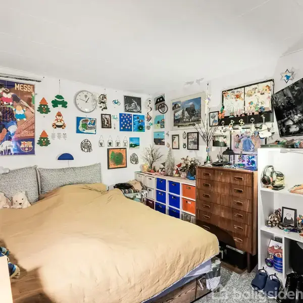 Soveværelse med en dobbeltseng dækket af en beige dyne. Omgivet af farverige plakater og dekorationer på væggene. En kommode og opbevaringskasser ved siden af sengen. TV hænger på væggen.