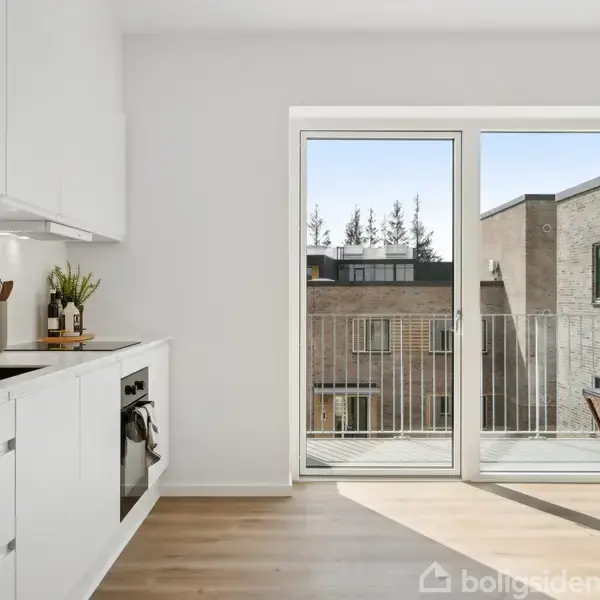 Et moderne køkken med hvide skabe og indbygget ovn strækker sig langs venstre væg. Der er en åben glasdør til en altan med udsigt over nabobygninger.