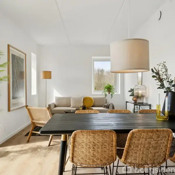 Et stilfuldt spisebord med flettede stole står i en moderne stue. En sofa og lamper skaber en hyggelig atmosfære, mens potteplanter tilføjer naturlig dekoration.