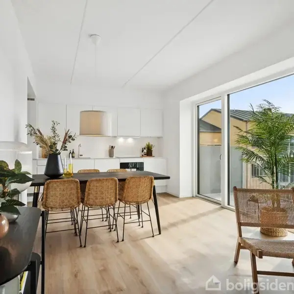 Et moderne køkken med en lille spiseplads. Bordet er omgivet af flettede stole. Store vinduer giver udsigt til en terrasse. Rummet er lyst, dekoreret med planter.
