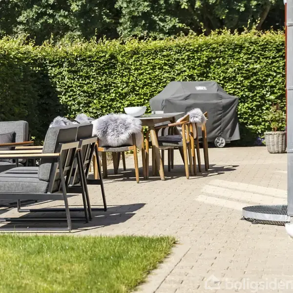 Udendørs terrasse med moderne møbler, hvor et bord er dækket med fåreskind på stolene. En gasgrill står i baggrunden på en flisebelagt overflade, omgivet af grønne buske.